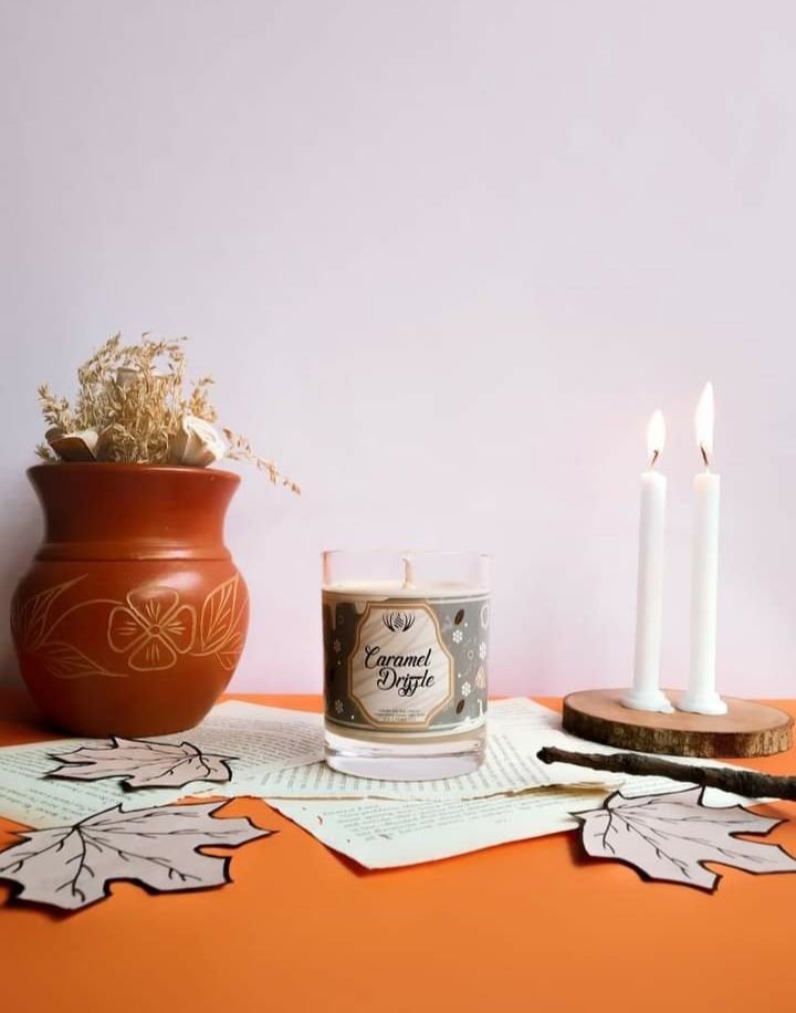 Caramel Drizzle candle: a grey and cream label with an arched frame, styled with a terracotta pot and paper leaves on orange.